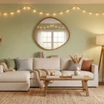 Cozy living room with light green walls, warm string lights, a beige sectional sofa, asymmetrical wooden mirror, tripod floor lamp, and natural pampas grass decor.